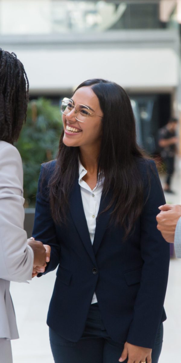 Female diverse business partners shaking hands with each other in hallway. Business man and women standing in office hall, talking and smiling. Successful partnership concept